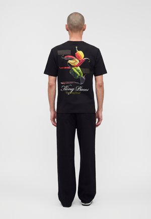 Bărbat purtând un tricou negru cu ardei colorați și textul „Filing Pieces Breaking Bread” pe spate, asortat cu pantaloni negri.