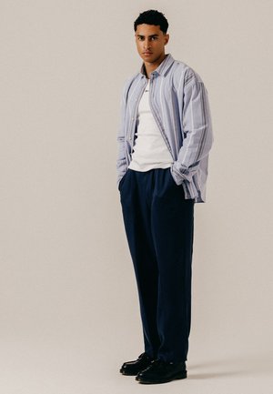 Jeune homme debout, les mains dans les poches, portant une chemise à rayures bleu clair sur un haut blanc, un pantalon bleu marine foncé et des chaussures noires sur fond uni.