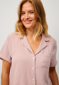 Světle růžová košile s krátkým rukávem vyrobená z texturované tkaniny, s límečkem, zapínáním na knoflíky a jednou kapsou na levé části hrudi.