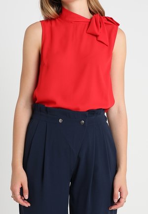 Femme portant un chemisier rouge sans manches avec un détail nœud à l'épaule et un pantalon bleu marine taille haute avec des boutons.