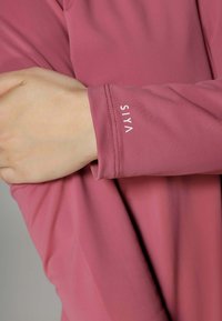 Langarmtop van zacht roze stof met een gladde textuur. Voorzien van een strakke manchette met de geperste merknaam "SIYA" in het wit.