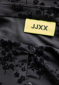 Melna auduma ar ziedu tekstūru, ar dzeltenu birku ar uzrakstu "JJXX" un melnu pogu. Gluda, mīksta materiāls ar sarežģīto detaļu.