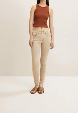 Kobieta ubrana w brązowy bezrękawnik typu crop top, beżowe obcisłe jeansy z podwójnym zapięciem na guziki oraz brązowe płaskie sandały z odkrytymi palcami, stojąca na jednolitym tle.