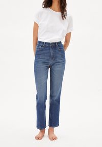 Person som bär högmidjade blå jeans och en vit t-shirt med kort ärm, står barfota mot en enkel vit bakgrund.