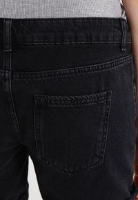 Zwarte spijkerbroek met een achterzak. De stof heeft een subtiele textuur, met zichtbare stiksels langs de naden en de randen van de zak.