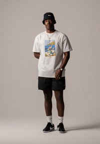 T-shirt en coton blanc avec un motif graphique coloré, shorts noirs, chaussures noires et un chapeau seau noir. Les accessoires comprennent des colliers et des chaussettes.