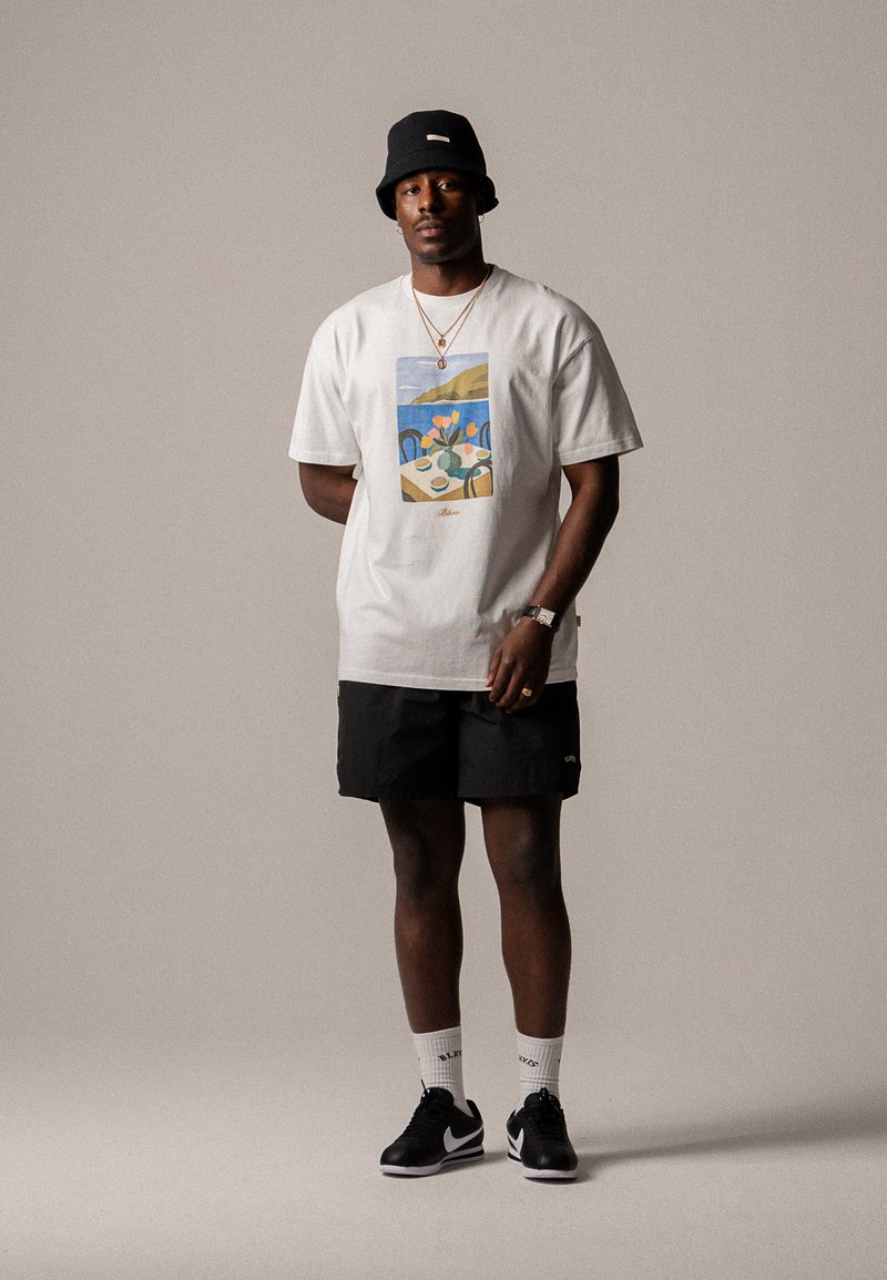 T-shirt en coton blanc avec un motif graphique coloré, shorts noirs, chaussures noires et un chapeau seau noir. Les accessoires comprennent des colliers et des chaussettes.