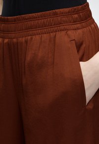 Gros plan d'une personne portant un pantalon marron à taille élastique avec une main partiellement dans la poche latérale sur un fond de haut noir.