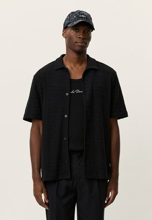 Zwart overhemdb met korte mouwen en een gestructureerd patroon, open kraag, knopen aan de voorkant, gedragen over een zwart tanktop, gecombineerd met een donker patroonpetje.