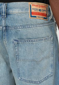Jean en denim bleu clair avec une étiquette rectangulaire marron sur la ceinture. Il comporte une poche arrière avec un design cousu.