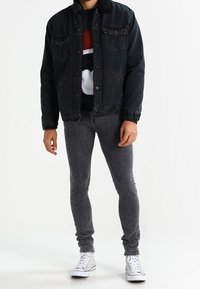 Homme portant une veste en denim noire avec col en sherpa, jean skinny gris foncé, pull graphique et baskets blanches montantes, debout sur un sol blanc.