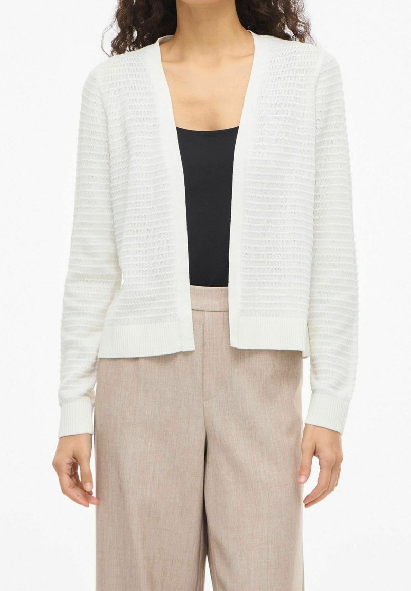 Femme portant un cardigan blanc texturé sur un haut noir et un pantalon beige taille haute, debout les bras détendus le long du corps.
