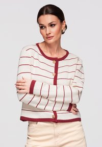 Femme aux cheveux foncés coiffés en chignon bas, portant un pull crème à rayures bordeaux et des boutons, bras croisés, regardant légèrement vers la droite sur un fond uni.