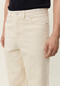 Bež manšestarske hlače rebraste teksture, s prednjim džepovima i kopčanjem na gumb. Dizajn je krojen za prilagođeni, uski izgled.