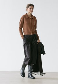 Vrouw lopend, gekleed in een bruin doorgestikt vest met knopen, zwarte brede broekspijpen, zwarte laarzen tot aan de knie, met een zwarte jas met franjes in haar hand.