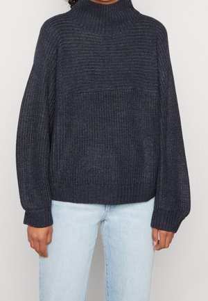 Person trägt einen dunkelgrauen, grob gestrickten Rollkragenpullover und hellblaue Jeans, steht vor einem schlichten Hintergrund.