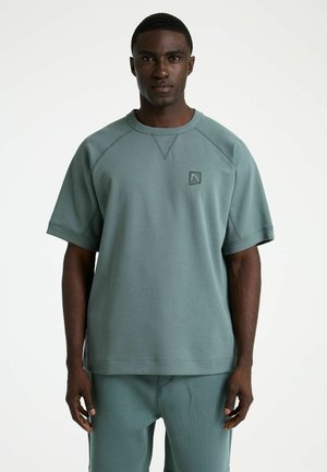 Man stands facing forward wearing loose-fitting muted teal short-sleeve sweatshirt and matching pants against plain white background.