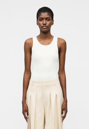 Γυναίκα με κοντά μαλλιά που φορά αμάνικο λευκό ριμπ πλεκτό τοπ και μπεζ υφασμάτινη φαρδιά παντελόνα με πιέτες, στέκεται μπροστά σε απλό φωτεινό φόντο.