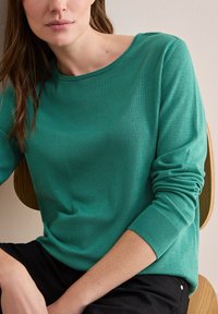 Grüner Strickpullover mit rundem Halsausschnitt und langen Ärmeln. Gerippte Bündchen und eine lockere Passform. Der Arm des Models ruht auf einem Holzstuhl.