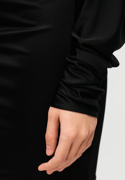 Μαύρο φόρεμα με μακριά μανίκια από απαλό, ελαστικό ύφασμα. Το μανίκι έχει λεπτομέρειες με πιέτες, τονίζοντας το εφαρμοστό σχέδιο.