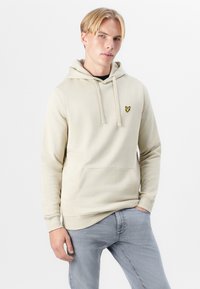 Hoodie beige en tissu doux, avec une poche kangourou et un logo de papillon jaune brodé sur la poitrine gauche.