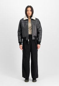 Veste bomber en cuir noir avec un col en shearling blanc, fermeture éclair, poignets côtelés, associée à un pantalon large noir et des bottes noires.