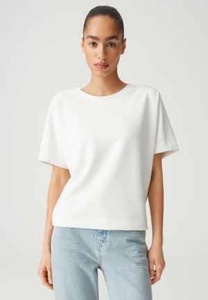 Vrouw met achterovergekamd haar, gekleed in een effen wit T-shirt met korte mouwen en lichtblauwe spijkerbroek, staand tegen een neutrale achtergrond.