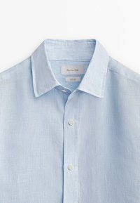 Hellblaues Leinenhemd mit strukturierter Webart, Knopfleiste und klassischem Kragen mit dezentem Streifenmuster. Etiketten sind innen sichtbar.