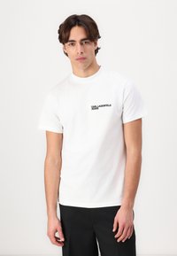 Weißes Baumwoll-T-Shirt mit kurzen Ärmeln und rundem Ausschnitt, mit einem schwarzen Logo-Text auf der linken Brustseite. Getragen mit schwarzen Hosen.