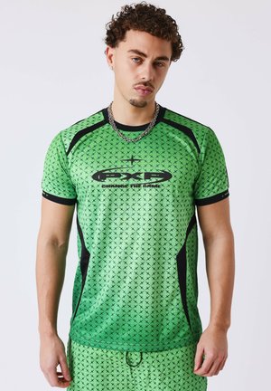 Jeune homme aux cheveux bouclés portant une tenue de sport verte avec des motifs en X noirs et des panneaux latéraux noirs, debout devant un fond uni.