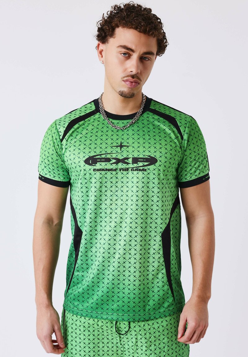 Jeune homme aux cheveux bouclés portant une tenue de sport verte avec des motifs en X noirs et des panneaux latéraux noirs, debout devant un fond uni.