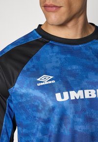 Sinine sportline särk mustade aktsentidega, millel on tekstuuriga muster, ümmargune kaelus ja valge "Umbro" logo ees.