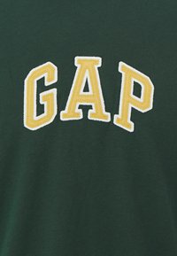 Tejido verde oscuro con grandes letras bordadas en amarillo y blanco que dicen "GAP" en un estilo colegial.