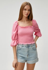 Lyserød smocket top med firkantet udskæring og korte puffærmer, parret med højtaljede denimshorts. Tekstileret stof og flæsekanter.