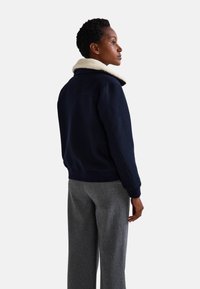 Veste de pilote de la marine avec un col en fausse fourrure crème, tissu texturé, poignets et ourlet côtelés. Portée avec un pantalon en laine gris.