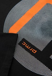 Černé tričko s kruhovým tečkovaným vzorem, oranžovým pruhem a textem "PMCM" v oranžové barvě na  černém látkovém pozadí.