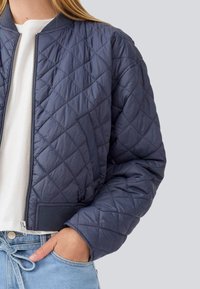 Veste bomber matelassée en bleu marine foncé, avec fermeture éclair sur le devant, poignets côtelés et finition texturée. Portée sur une chemise blanche.