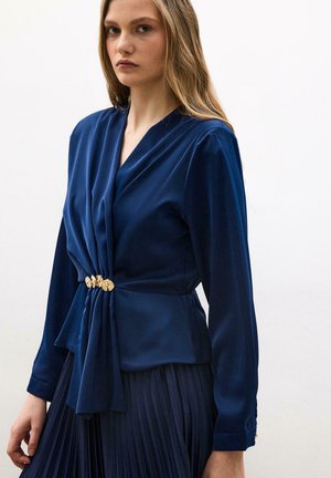 Jeune femme portant un chemisier bleu marine à manches longues froncées avec une boucle décorative dorée et une jupe plissée assortie, sur un fond uni.