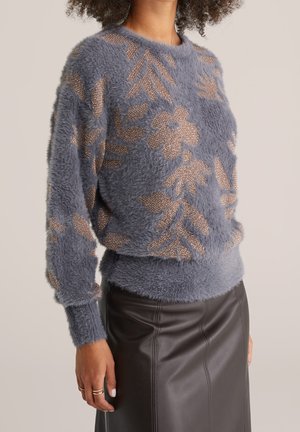 Modèle portant un pull duveteux bleu-gris à motifs floraux métallisés et une jupe marron foncé imitation cuir.