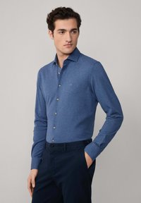 Hombre vestido con una camisa azul ajustada de botones, de manga larga, y pantalones navy, de pie con una mano en el bolsillo, frente a un fondo liso.
