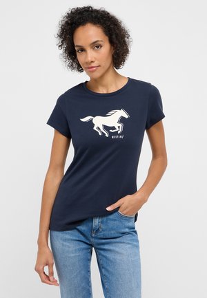 Frau mit kurzen lockigen Haaren, die ein marineblaues T-Shirt mit einem weißen Laufpferd-Motiv und hellblaue Jeans trägt, die Hand in der Tasche.