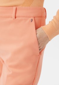 Peach-colored trousers made of textured fabric, featuring a side pocket and a small metallic logo accent on the waistband.