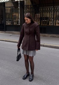 Hnedý elegantný kabát s vysokým golierom, šedá plisovaná sukňa, čierne pančuchy a mokasíny. Čierna kabelka dokončuje outfit.