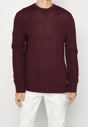 Mand iført en langærmet, mørkerød crew neck sweater kombineret med hvide bukser, stående mod en ensfarvet lys baggrund.