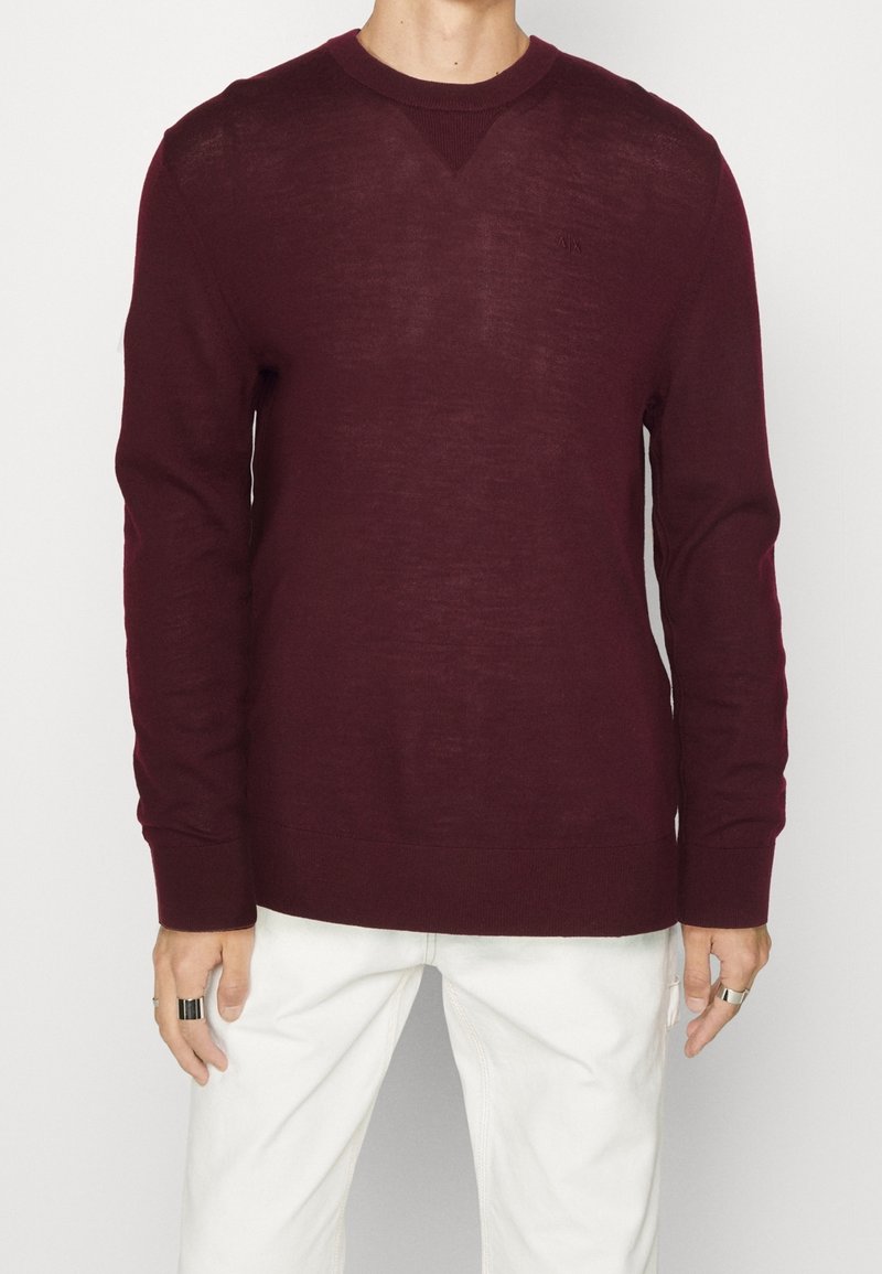 Homme portant un pull bordeaux à col rond et manches longues, associé à un pantalon blanc, debout devant un fond clair uni.