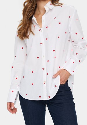 Donna che indossa una camicia bianca con bottoni e motivo a cuori rossi, abbinata a jeans blu scuro, mano in tasca e maniche larghe.