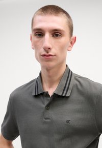 Šedé polo tričko s texturovaným povrchem, s dvoutlačítkovou légou a pruhovaným límcem v tmavších odstínech. Vyšité logo na hrudi.