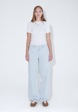 Jonge vrouw met lang blond haar staat tegen een witte achtergrond, draagt een wit kortemouwtopje, lichtblauwe wijde jeans en bruine sneakers.