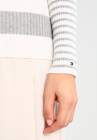 Maglione a costine con strisce grigie e bianche, caratterizzato da un piccolo logo del marchio ricamato sul polsino. Texture morbida e design aderente.