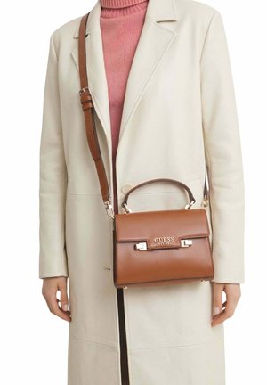 Femme portant un manteau crème et un col roulé rose, tenant un sac à main Guess en cuir marron avec poignée supérieure et bandoulière ajustable.